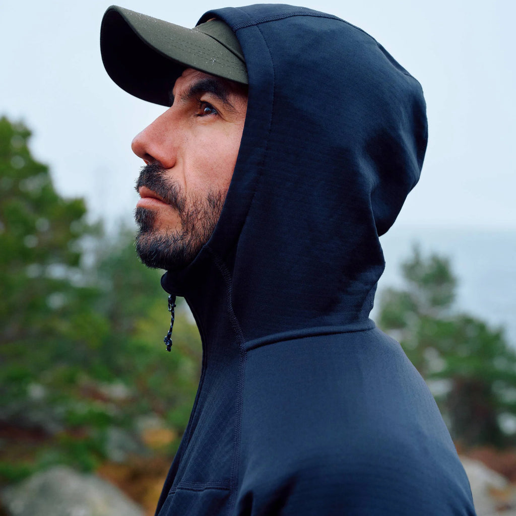 Man wearing a dark blue hooded jacket and cap in a natural setting with trees and rocks.