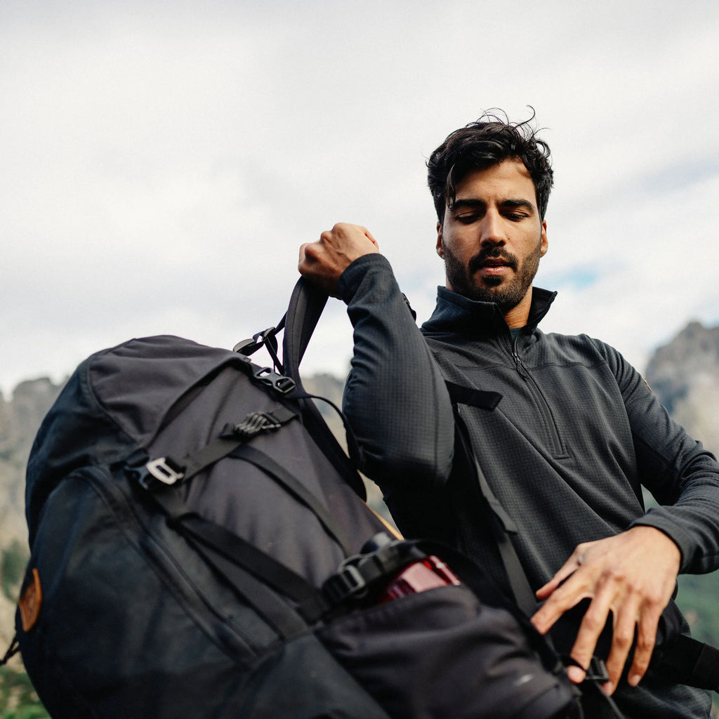 Man wearing a black fleece holding a black backpack with mountains in the background
